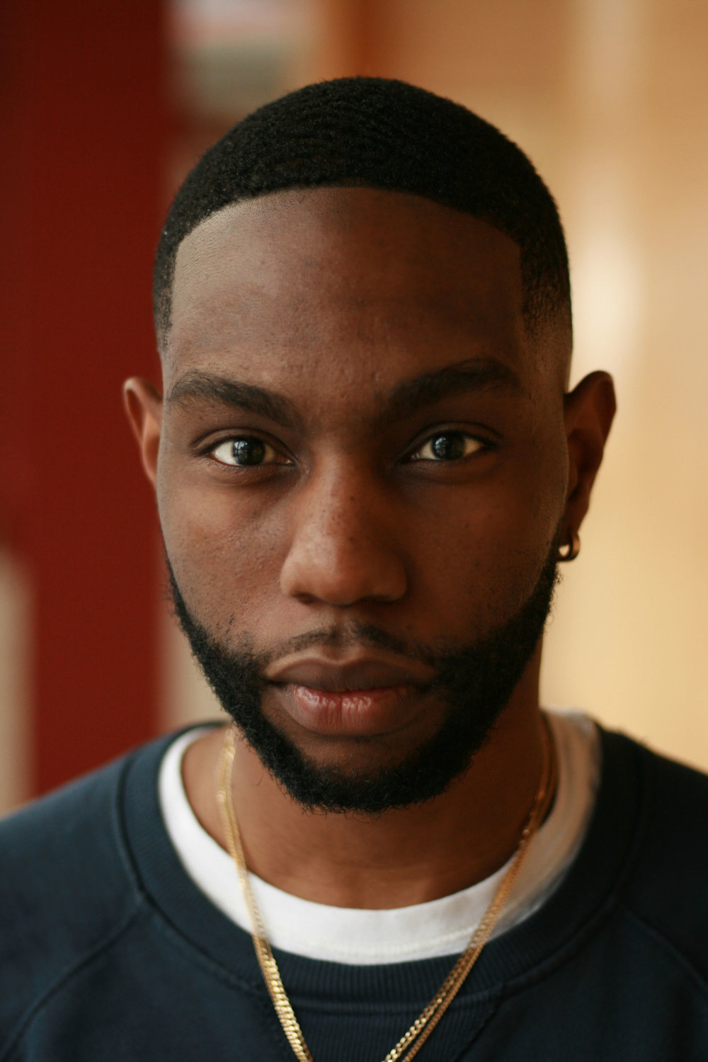 A close up portrait of Steven with the background out of focus. He has a gold hoop earing in his left ear, is wearing two gold chains and a black sweater.
